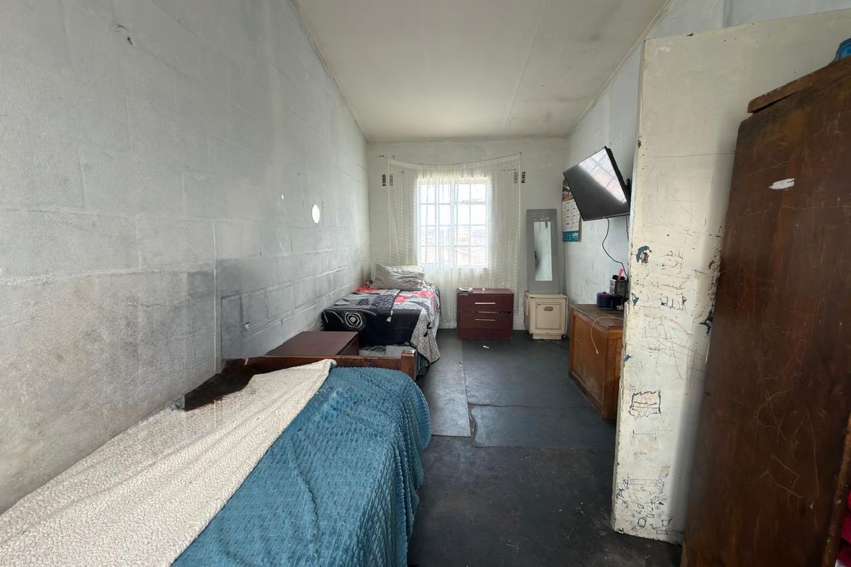 A bedroom featuring exposed concrete block walls, a window with security bars, and worn concrete flooring, requiring cosmetic updates and refurbishment.