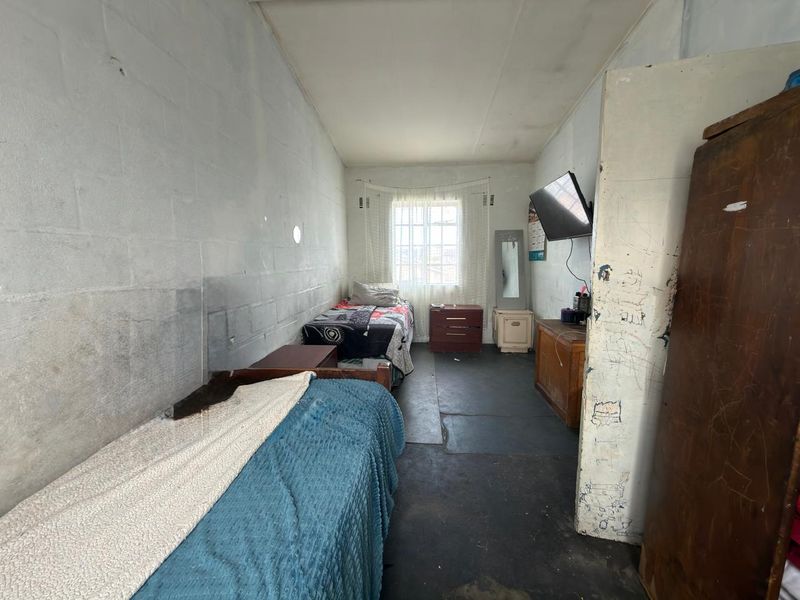 A bedroom featuring exposed concrete block walls, a window with security bars, and worn concrete flooring, requiring cosmetic updates and refurbishment.