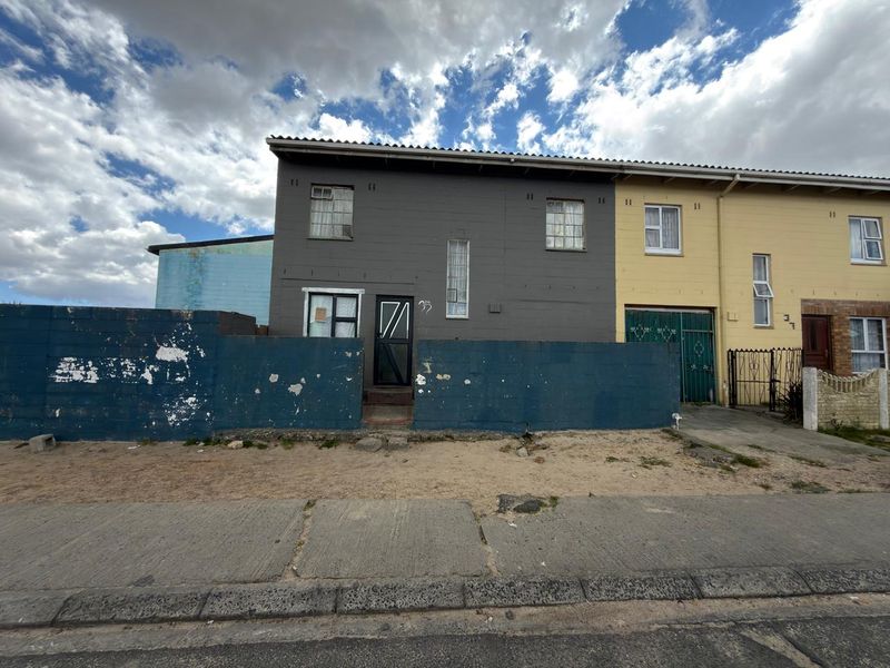 Front elevation of a two-story townhouse featuring a dark grey facade, security gate, and a weathered blue boundary wall requiring maintenance.