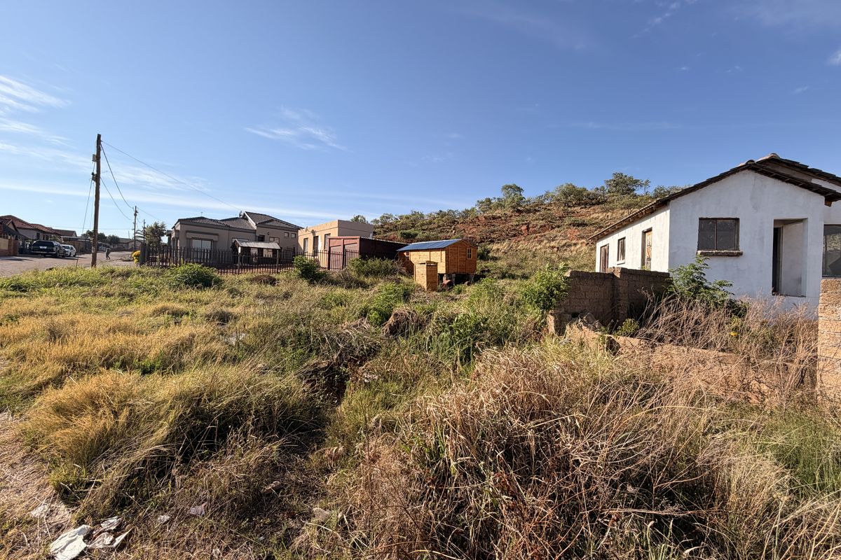 A view of a residential vacant land plot featuring overgrown vegetation, a boundary wall, and proximity to existing neighborhood infrastructure.