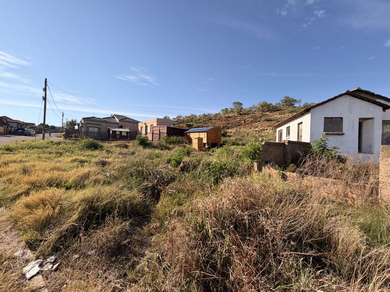 A view of a residential vacant land plot featuring overgrown vegetation, a boundary wall, and proximity to existing neighborhood infrastructure.