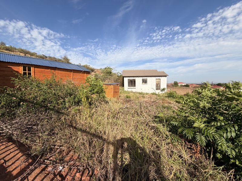 A view of a residential plot featuring a timber cabin and a small concrete structure set within an overgrown garden area.