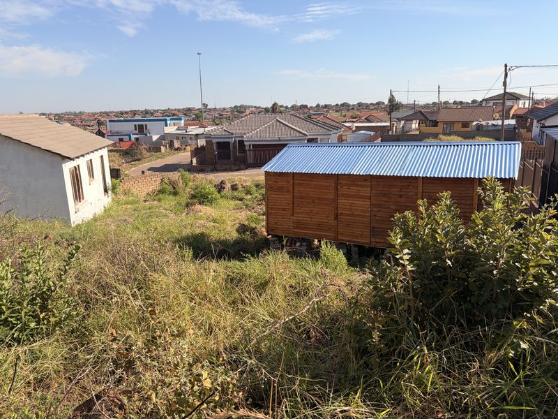 A view of a residential plot featuring a small timber structure with a corrugated metal roof and surrounding overgrown vegetation.
