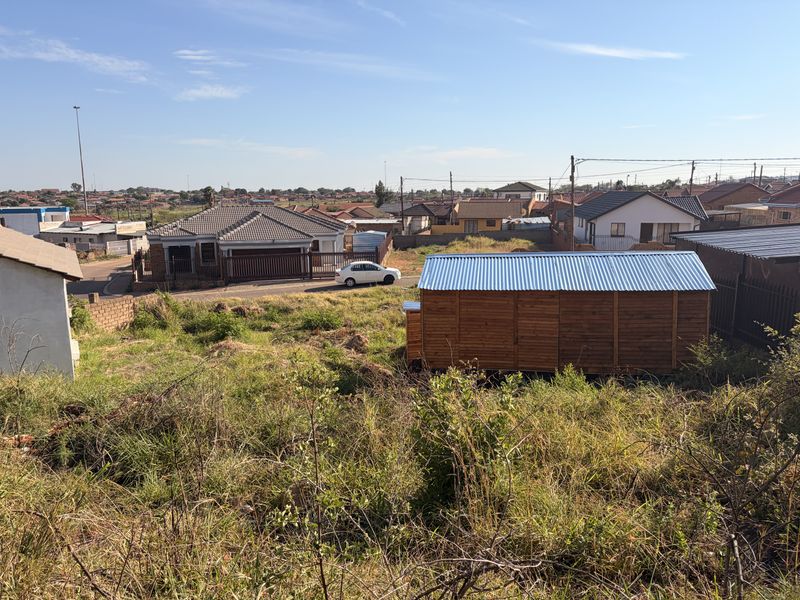 A view of a residential plot featuring overgrown vegetation and a small timber structure with a corrugated metal roof.