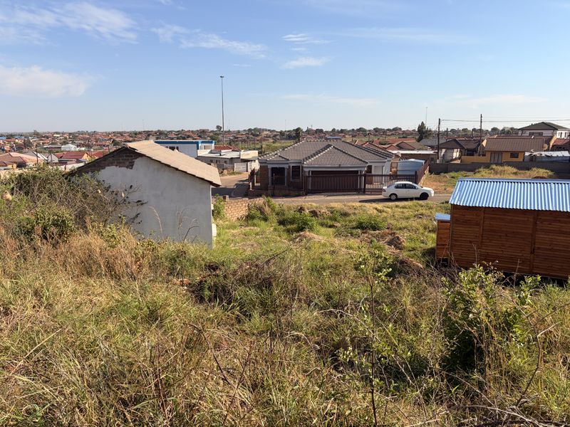 An elevated view of a vacant residential plot featuring overgrown grass, a small outbuilding, and a corrugated metal shed in a suburban neighborhood.