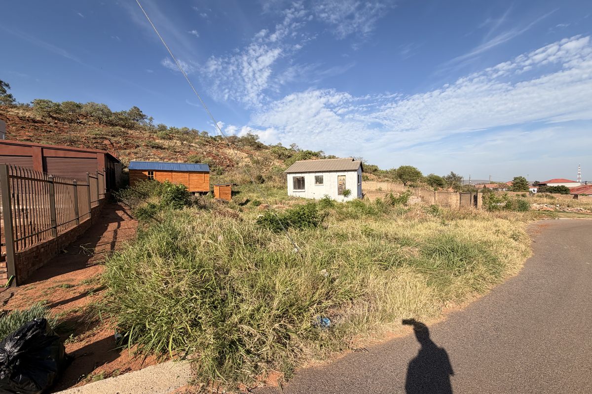 A vacant residential plot featuring a small outbuilding, boundary wall, and direct access to a paved road, set against a hillside landscape.