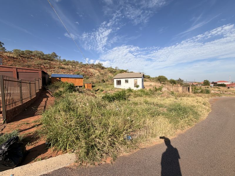 A vacant residential plot featuring a small outbuilding, boundary wall, and direct access to a paved road, set against a hillside landscape.