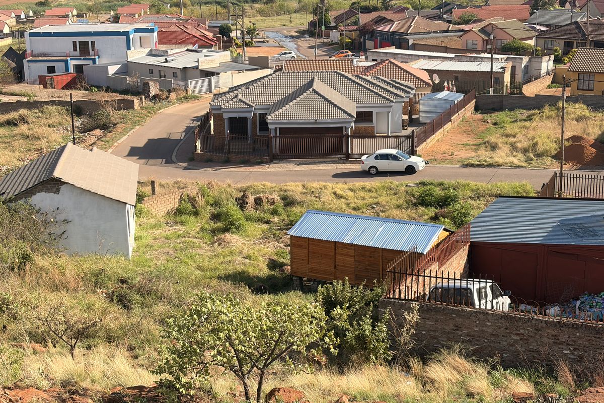 Aerial view of a residential plot featuring a vacant sloped area, perimeter fencing, and surrounding neighborhood infrastructure.