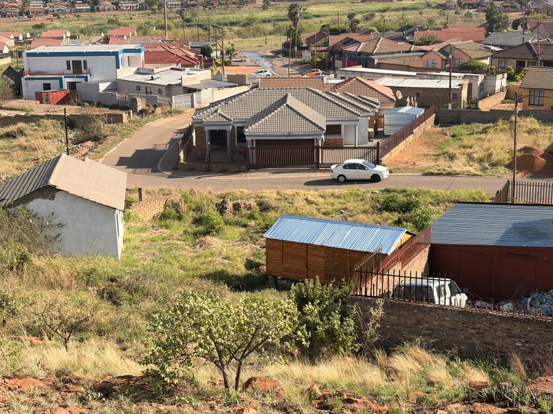 Aerial view of a residential plot featuring a vacant sloped area, perimeter fencing, and surrounding neighborhood infrastructure.