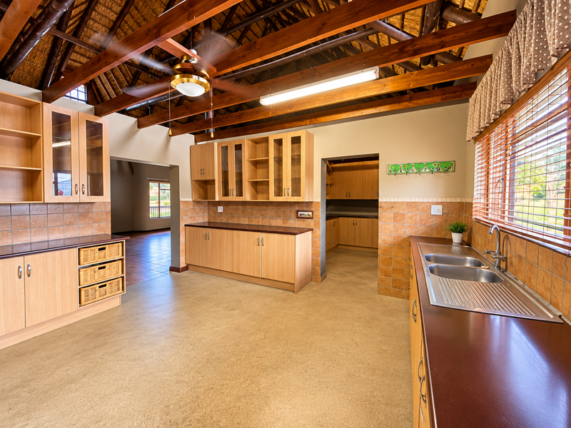 Spacious kitchen featuring exposed timber rafters, a thatched ceiling, extensive built-in cabinetry, and a double stainless steel sink with tiled splashbacks.