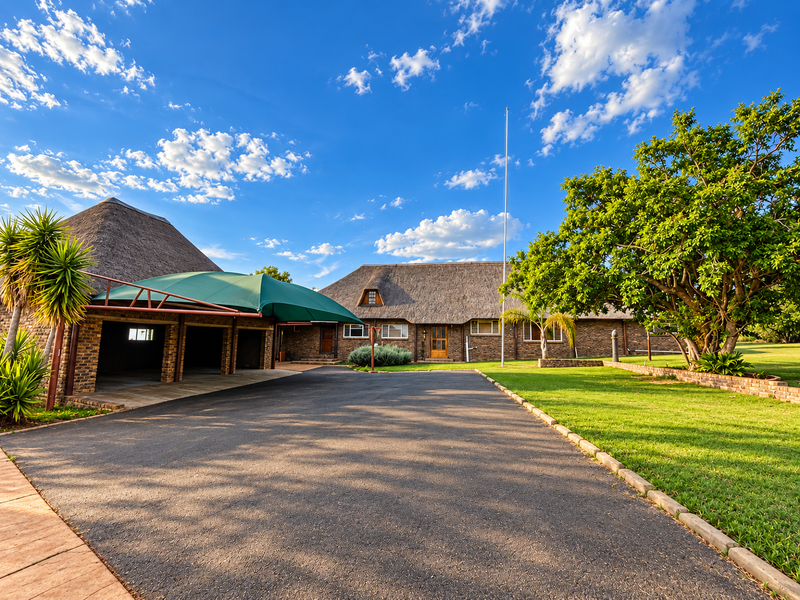 Spacious front exterior featuring a large asphalt driveway, a thatched roof house, and a shaded carport area set within a well-maintained garden.