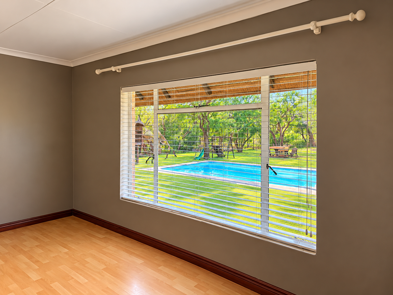 A bright, empty bedroom featuring laminate flooring, neutral walls, and a large window with venetian blinds overlooking the garden and swimming pool area.