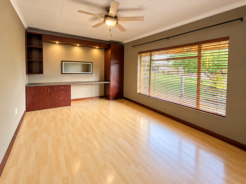 A bright, empty home office featuring built-in cabinetry, a desk, laminate flooring, and large windows with venetian blinds.