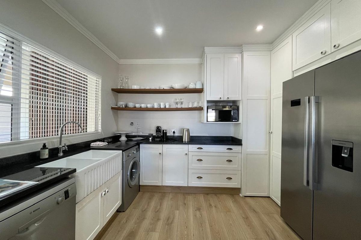 Modern kitchen featuring white shaker cabinetry, black granite countertops, a farmhouse sink, open timber shelving, and integrated appliances with light wood-look flooring.