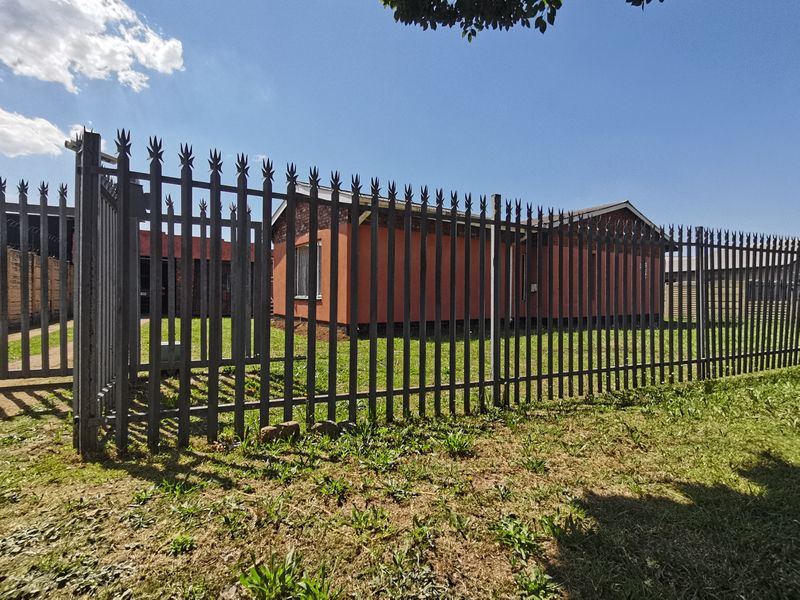 A view of the property's front exterior, featuring a secure metal perimeter fence and a brick-walled house with a tiled roof.