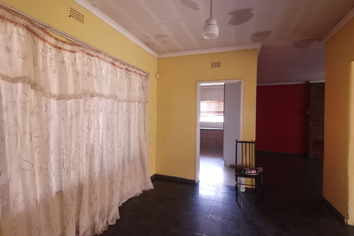 Spacious living area featuring dark tiled flooring and yellow walls, with visible ceiling maintenance required. Leads through to a kitchen area.