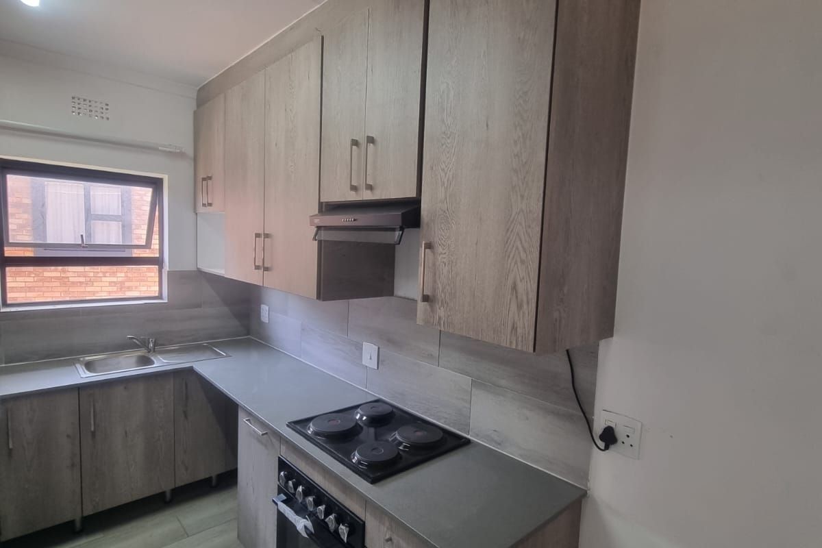 A clean, modern kitchen featuring wood-look cabinetry, integrated electric hob and oven, stainless steel sink, and neutral tiled flooring.