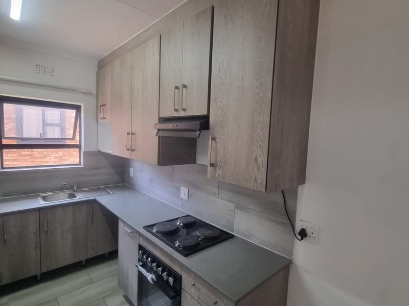 A clean, modern kitchen featuring wood-look cabinetry, integrated electric hob and oven, stainless steel sink, and neutral tiled flooring.