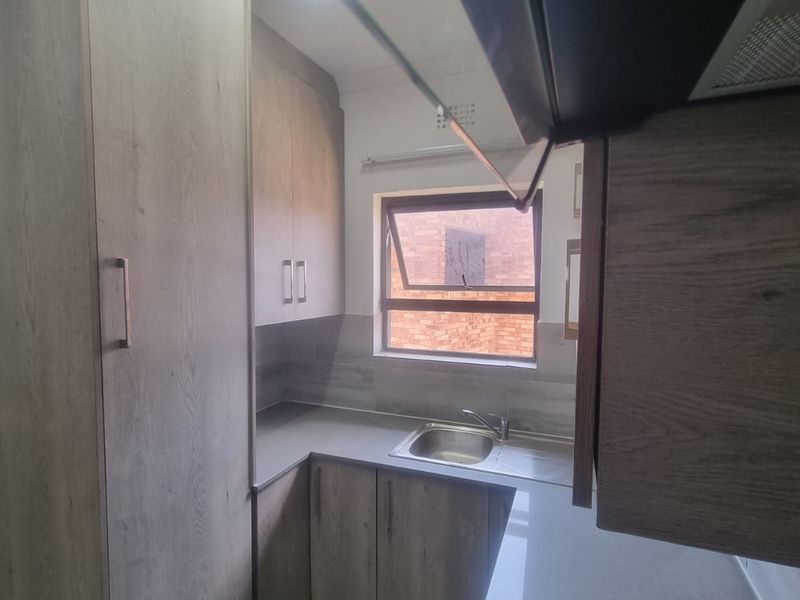 A modern, compact kitchen featuring light wood-grain cabinetry, stone countertops, a stainless steel sink, and tiled backsplash with natural light from a window.
