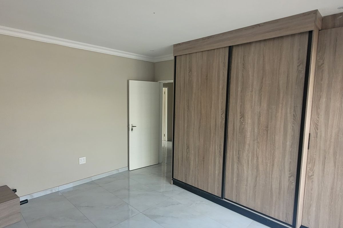 A bright, empty bedroom featuring modern wood-finish built-in cupboards, neutral wall tones, and polished floor tiles.
