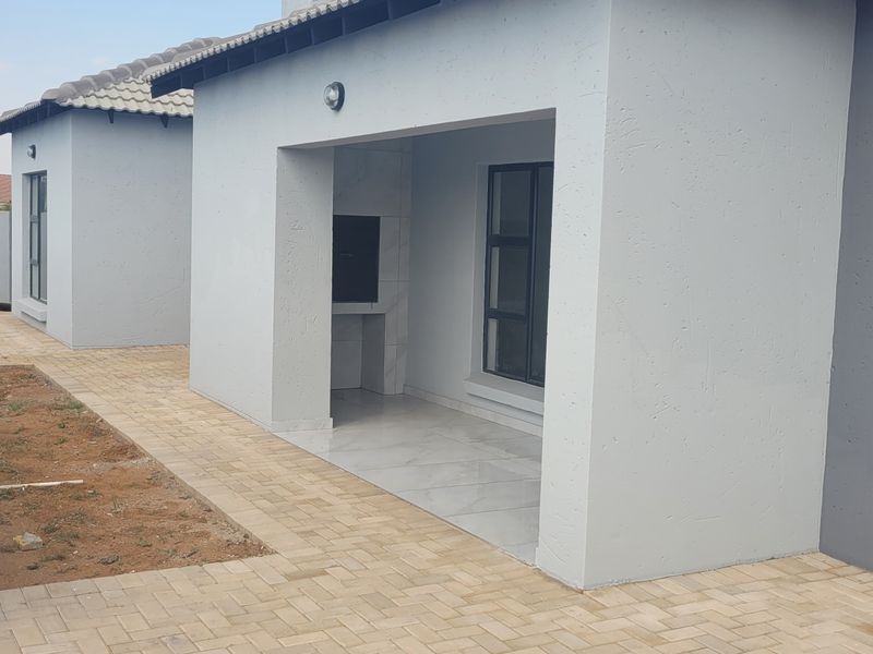 Rear view of the property featuring a covered patio with a built-in braai, paved walkways, and a garage, set against a landscaped boundary wall.