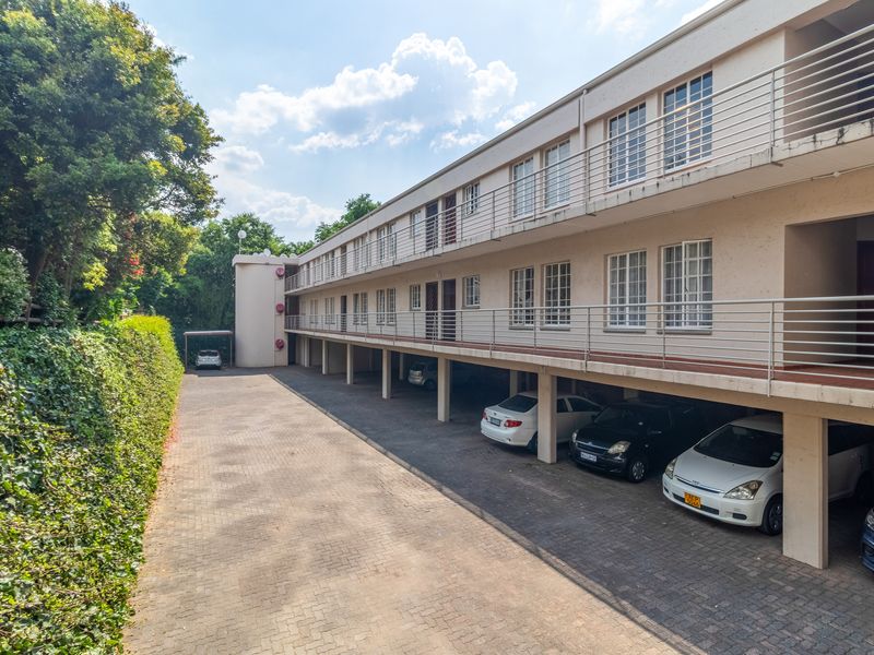 Exterior view of a multi-story apartment complex featuring covered ground-level parking bays, paved driveway, and balcony access for upper-level units.