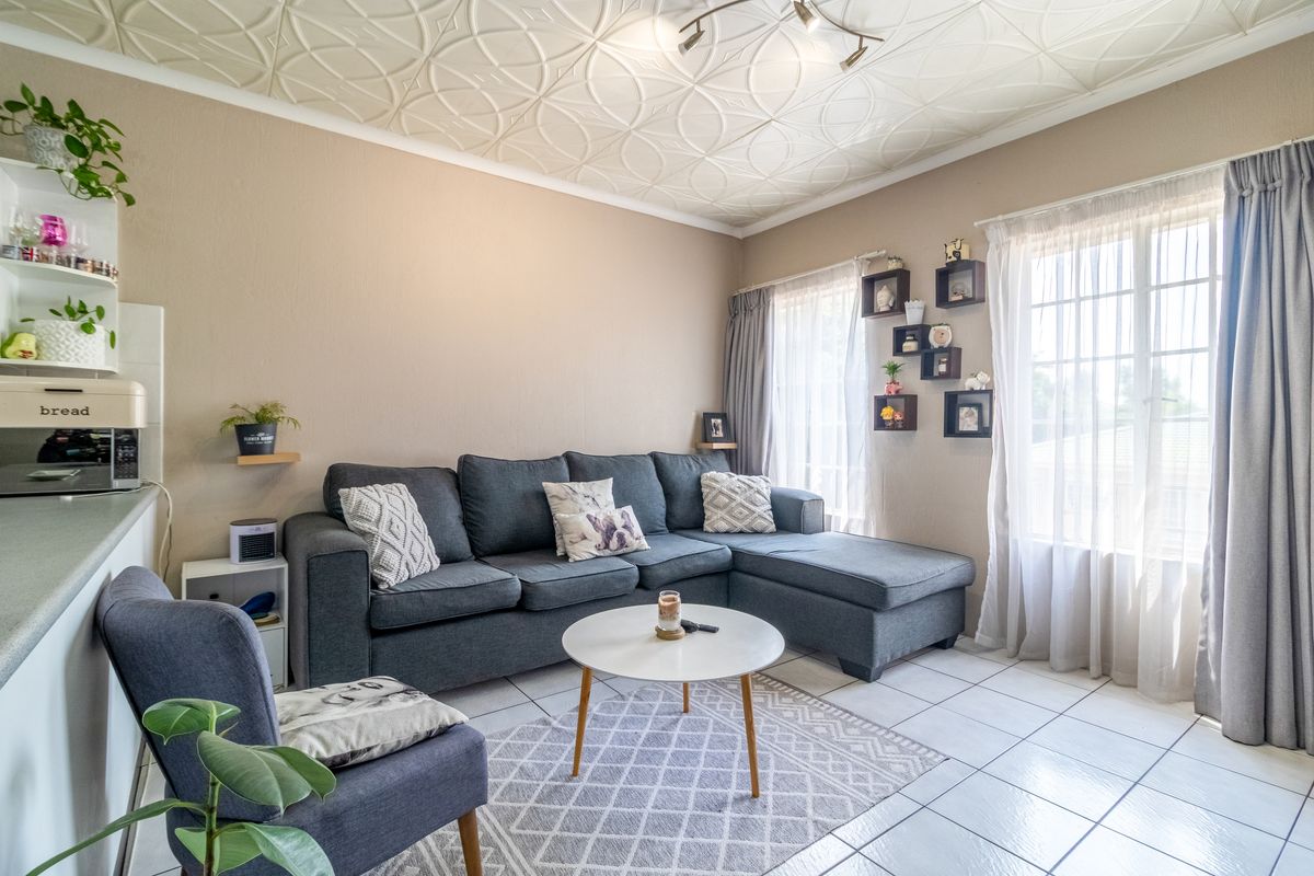 A comfortable living room featuring tiled flooring, a decorative patterned ceiling, and large windows with curtain rails, providing a bright and functional living space.