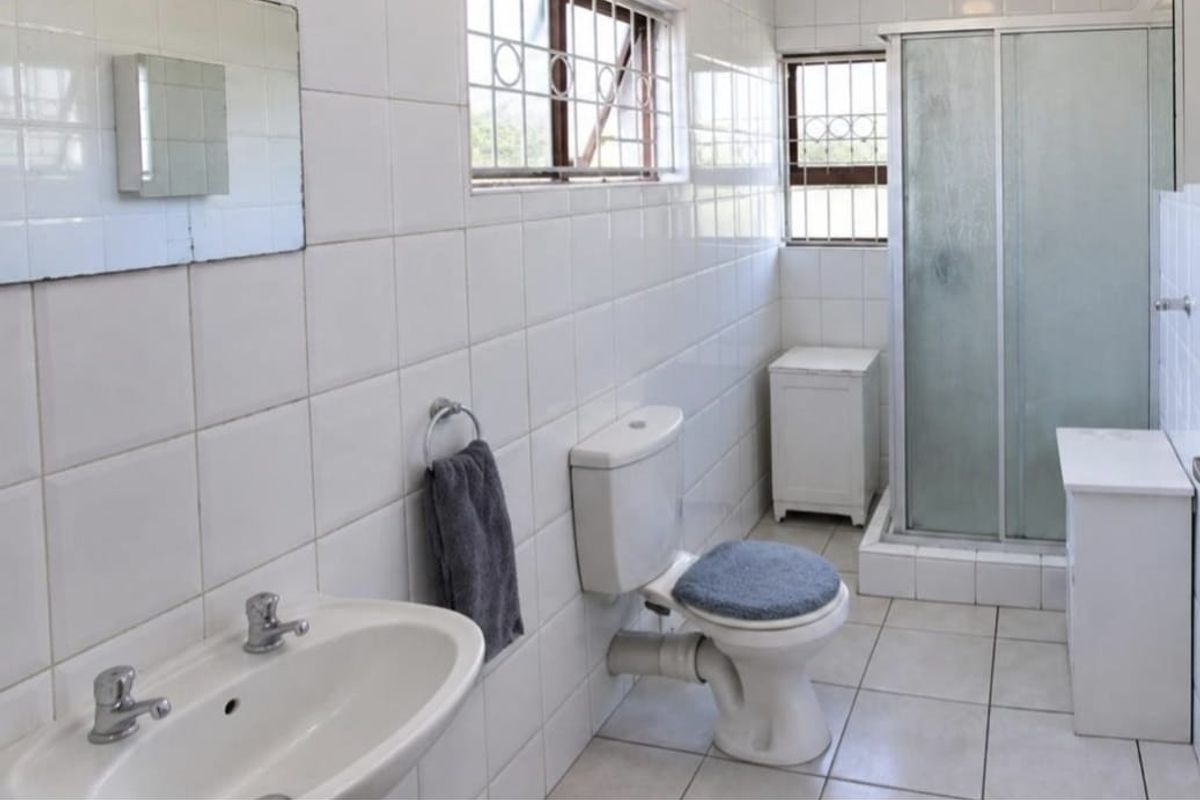 A functional bathroom featuring a shower enclosure, pedestal basin, and toilet, finished with white wall tiles and security bars on the windows.