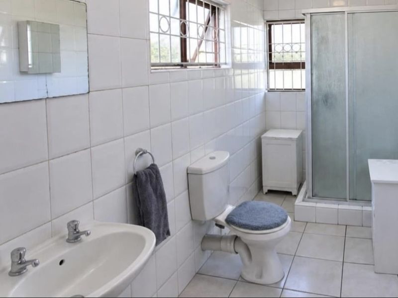 A functional bathroom featuring a shower enclosure, pedestal basin, and toilet, finished with white wall tiles and security bars on the windows.
