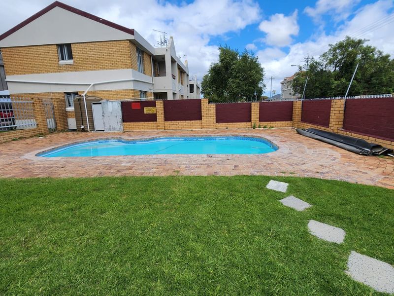 Communal swimming pool area featuring brick paving, a surrounding lawn, and a secure perimeter wall with security spikes.