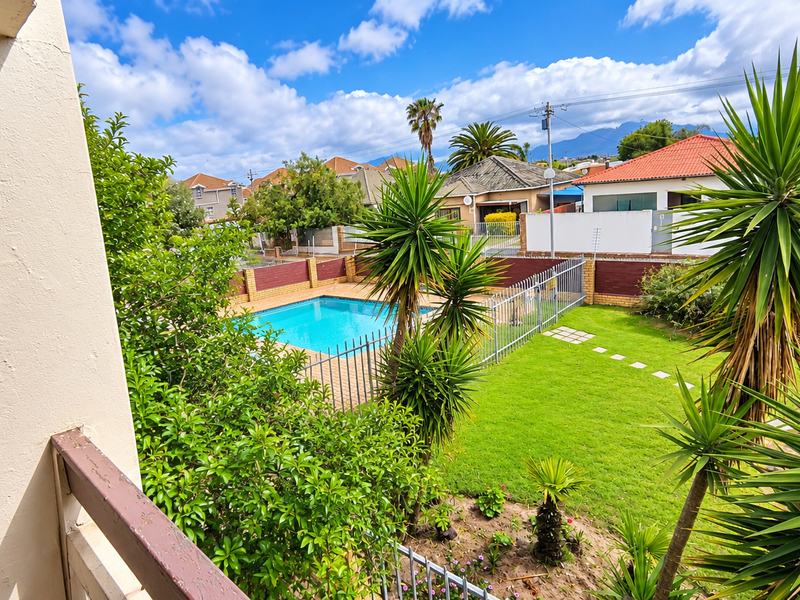 View of a communal swimming pool area with metal fencing, a well-maintained lawn, and established garden landscaping.