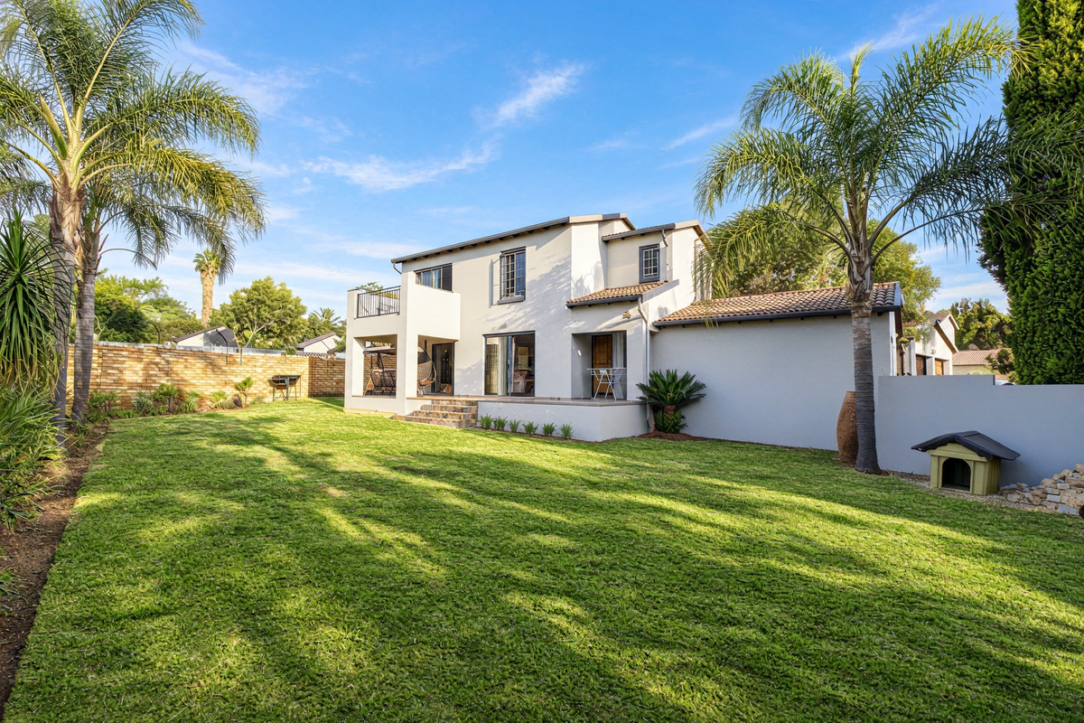 A well-maintained rear exterior view of the townhouse featuring a spacious lawn, a covered patio, and an upper-level balcony.