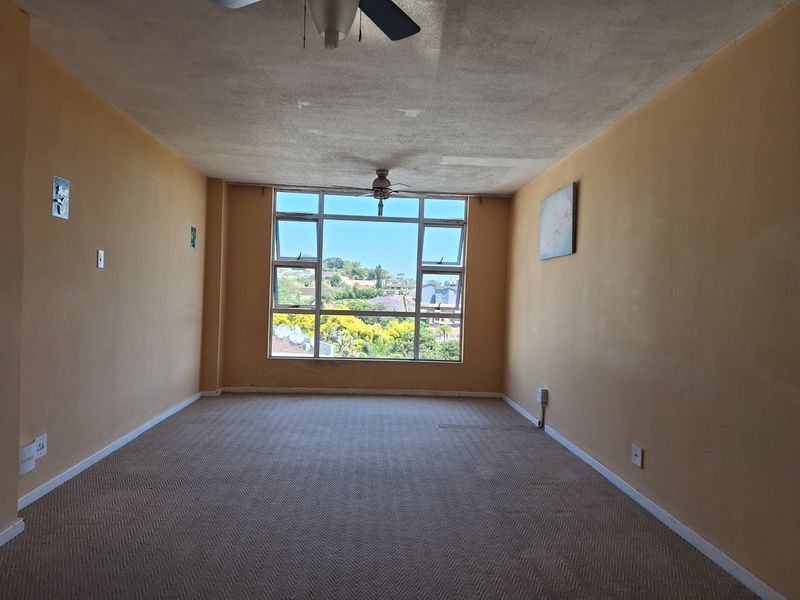An empty living room featuring carpeted flooring, a ceiling fan, and a large window providing natural light and neighborhood views.