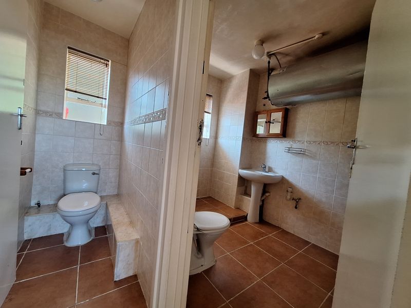A functional bathroom featuring tiled walls and floors, a pedestal basin, and a toilet, with an exposed geyser unit requiring aesthetic enclosure.