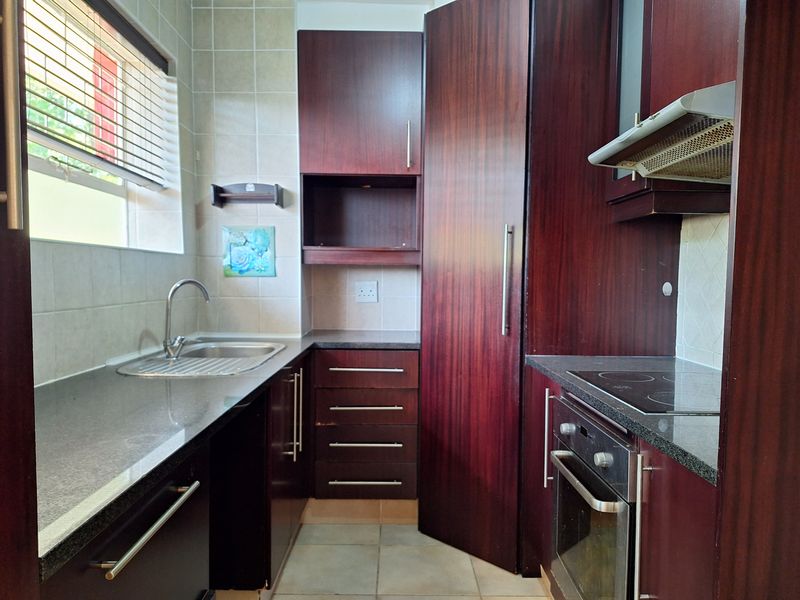 A compact kitchen featuring dark wood cabinetry, granite countertops, built-in oven, electric hob, and tiled finishes.