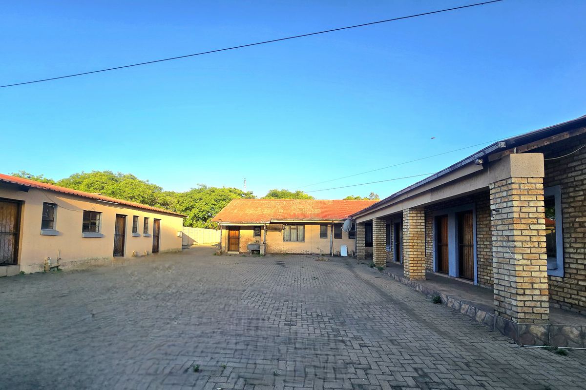 Rear exterior view of the property featuring a large paved courtyard, brick facade, and covered patio area with corrugated metal roofing.