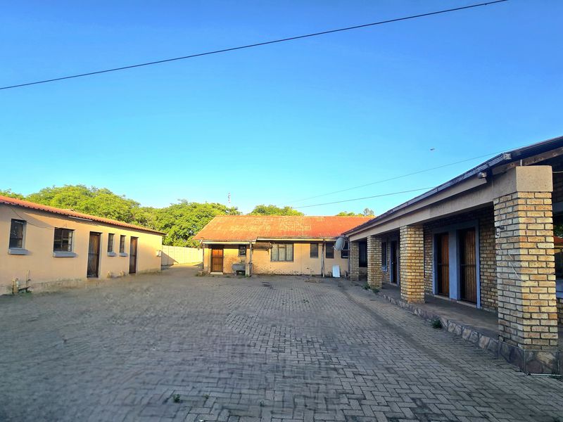 Rear exterior view of the property featuring a large paved courtyard, brick facade, and covered patio area with corrugated metal roofing.