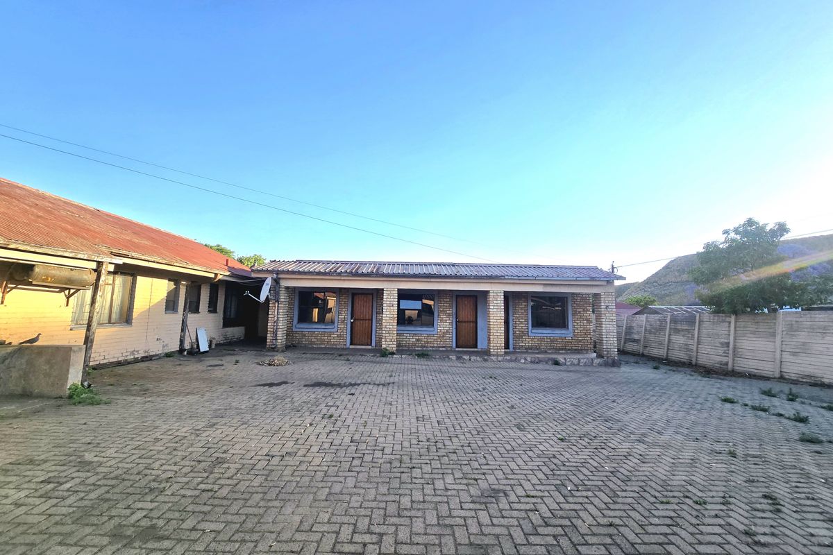 A rear exterior view of the property featuring a large paved courtyard, brick facade, and corrugated metal roofing with a surrounding boundary wall.