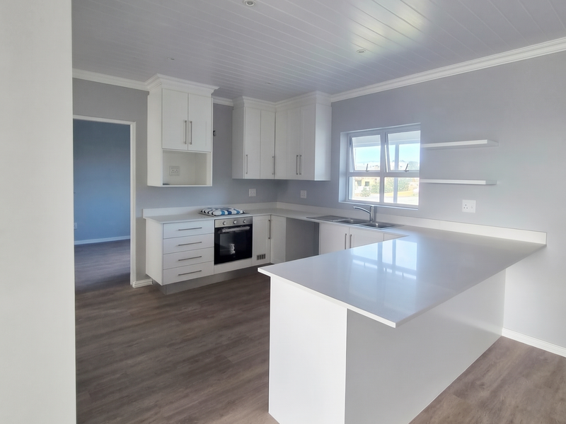 Modern, bright kitchen featuring white cabinetry, quartz countertops, integrated oven, and sleek laminate flooring in an open-plan layout.