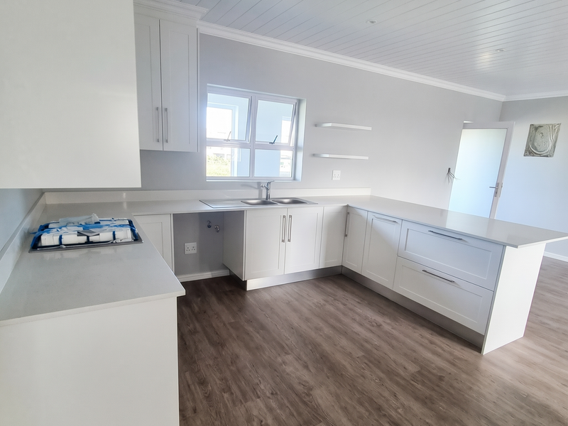 A modern, bright, and empty kitchen featuring white cabinetry, stone countertops, a stainless steel sink, and wood-look laminate flooring.