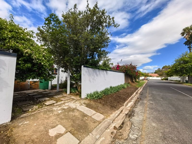 A street-level view of the property boundary, featuring a white perimeter wall, established trees, and a paved driveway entrance.