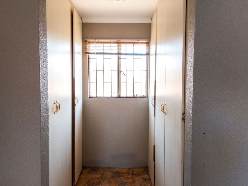 A functional bedroom corridor featuring built-in cupboards, tiled flooring, and a window fitted with security bars and venetian blinds.