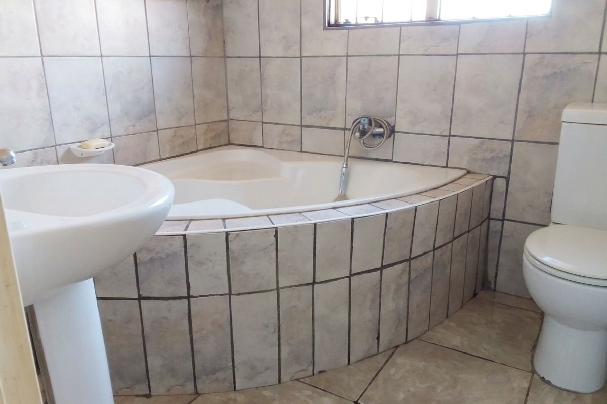 A dated bathroom featuring a corner bathtub, pedestal basin, and toilet with neutral wall and floor tiling.