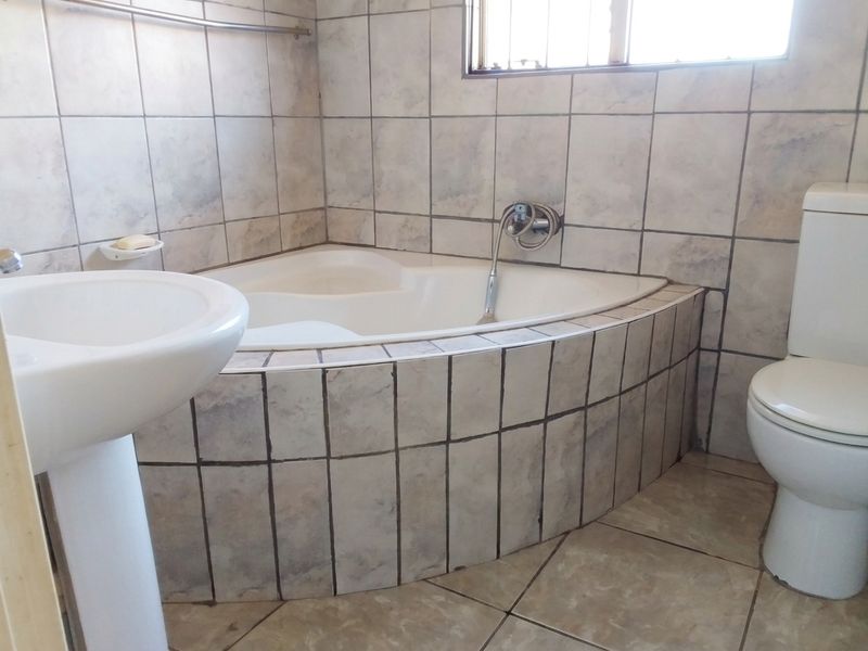A dated bathroom featuring a corner bathtub, pedestal basin, and toilet with neutral wall and floor tiling.