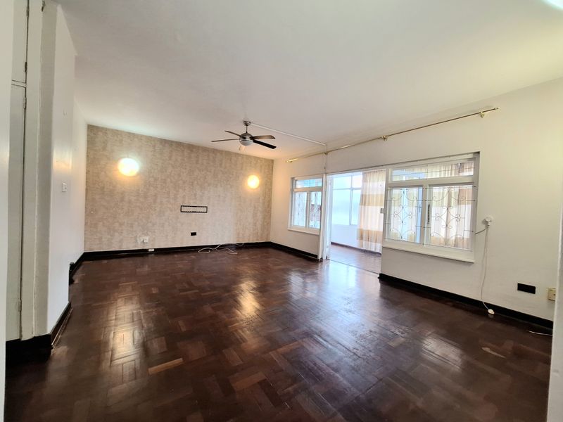 Spacious living area featuring classic parquet flooring, a ceiling fan, and large windows with security bars, leading to an adjoining enclosed balcony.
