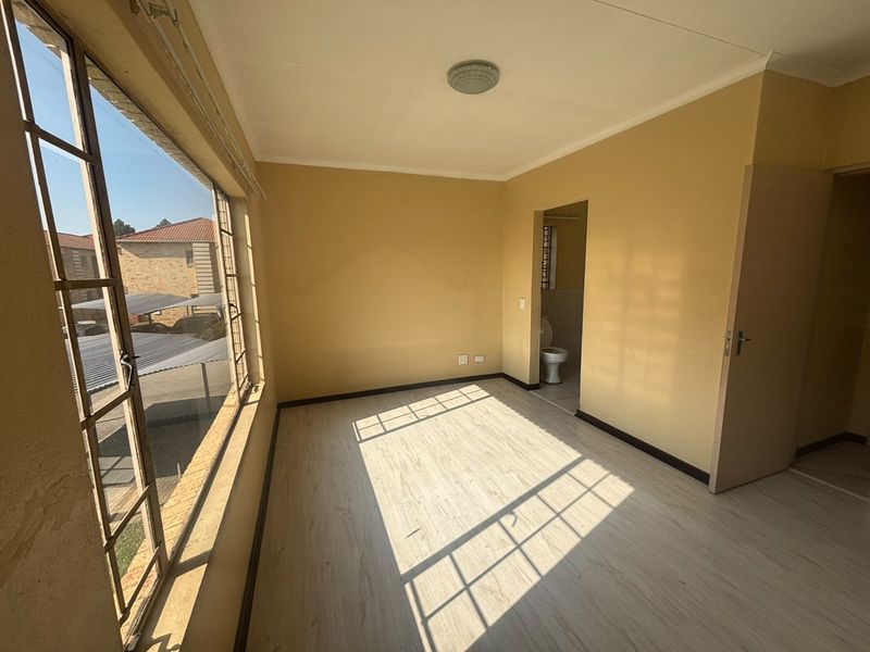 An empty bedroom featuring light-toned laminate flooring, large steel-framed windows, and an adjoining en-suite bathroom area.