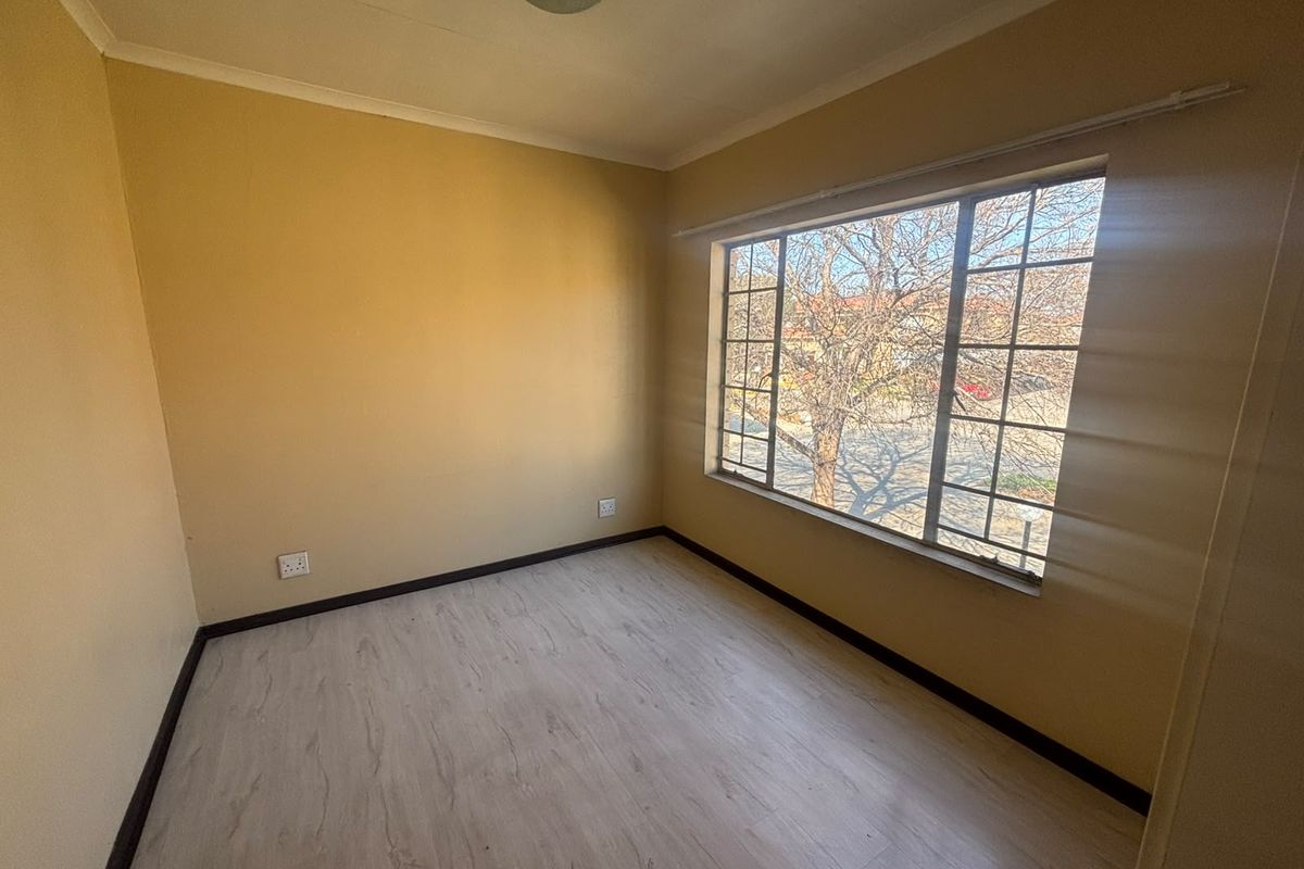 An empty bedroom featuring light-toned laminate flooring, a large window with security bars, and neutral wall paint, offering a blank canvas for personalization.