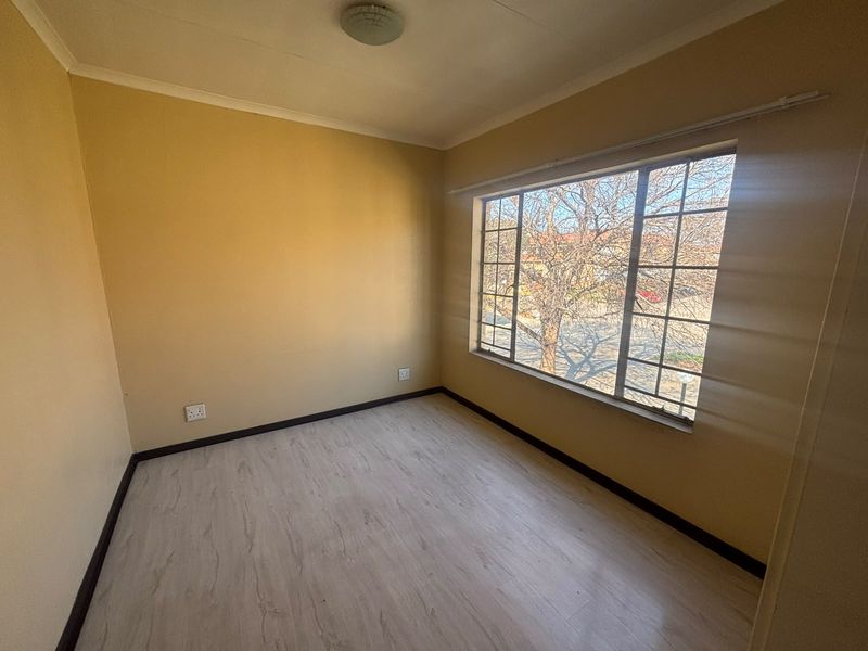 An empty bedroom featuring light-toned laminate flooring, a large window with security bars, and neutral wall paint, offering a blank canvas for personalization.