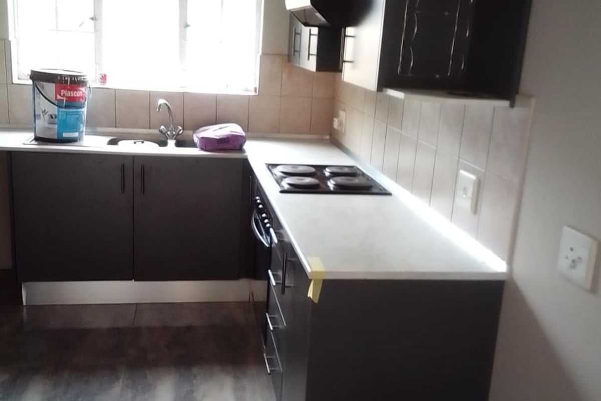 A compact kitchen featuring dark cabinetry, light countertops, an electric hob with extractor fan, and tiled flooring.