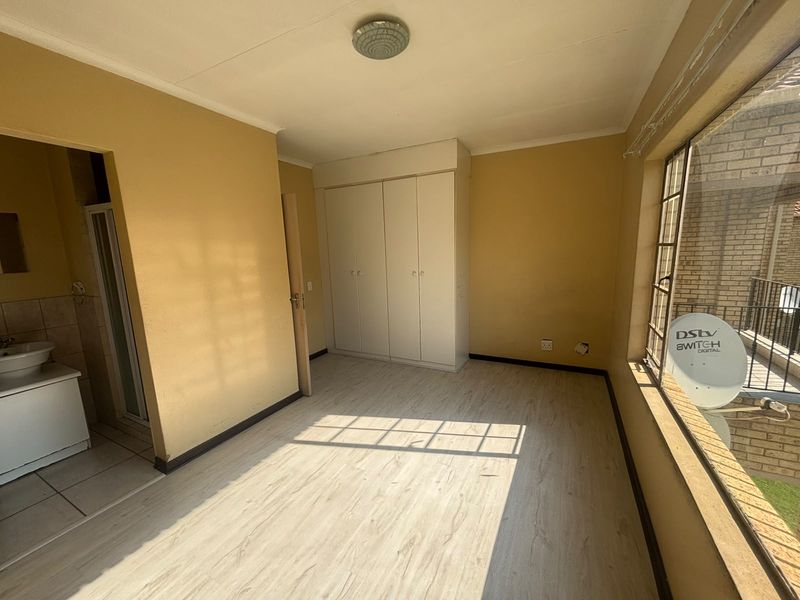 A bright, empty bedroom featuring laminate flooring, built-in cupboards, and large steel-framed windows, with an adjoining bathroom area.